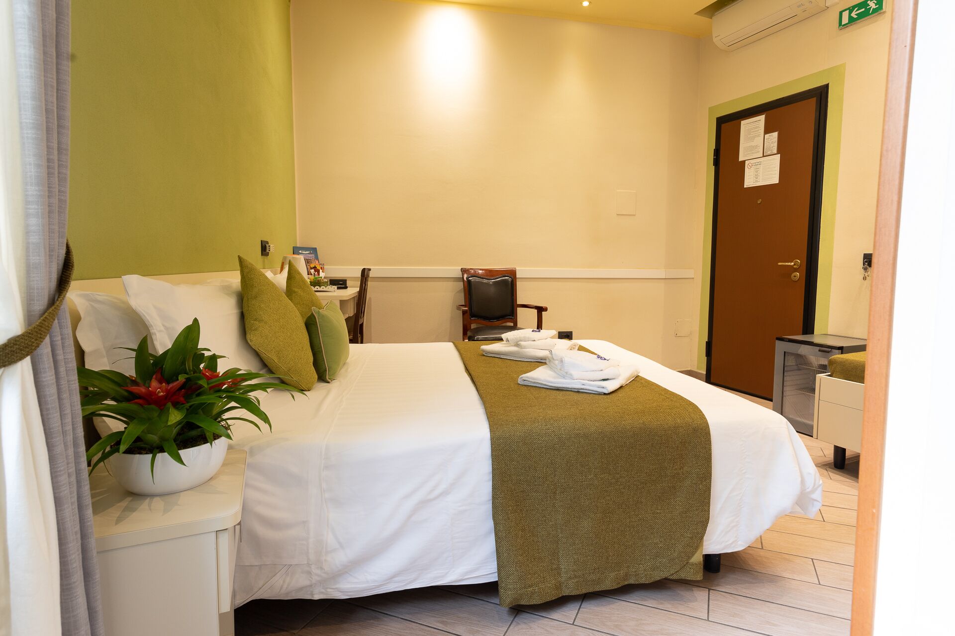 Una camera d'albergo con un letto, un comodino con una pianta, una sedia, una scrivania con un computer e una porta.