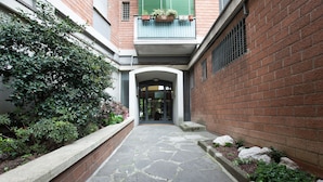 Un sendero de piedra que conduce a la entrada de un edificio, con un muro de ladrillo a un lado y un seto verde al otro.