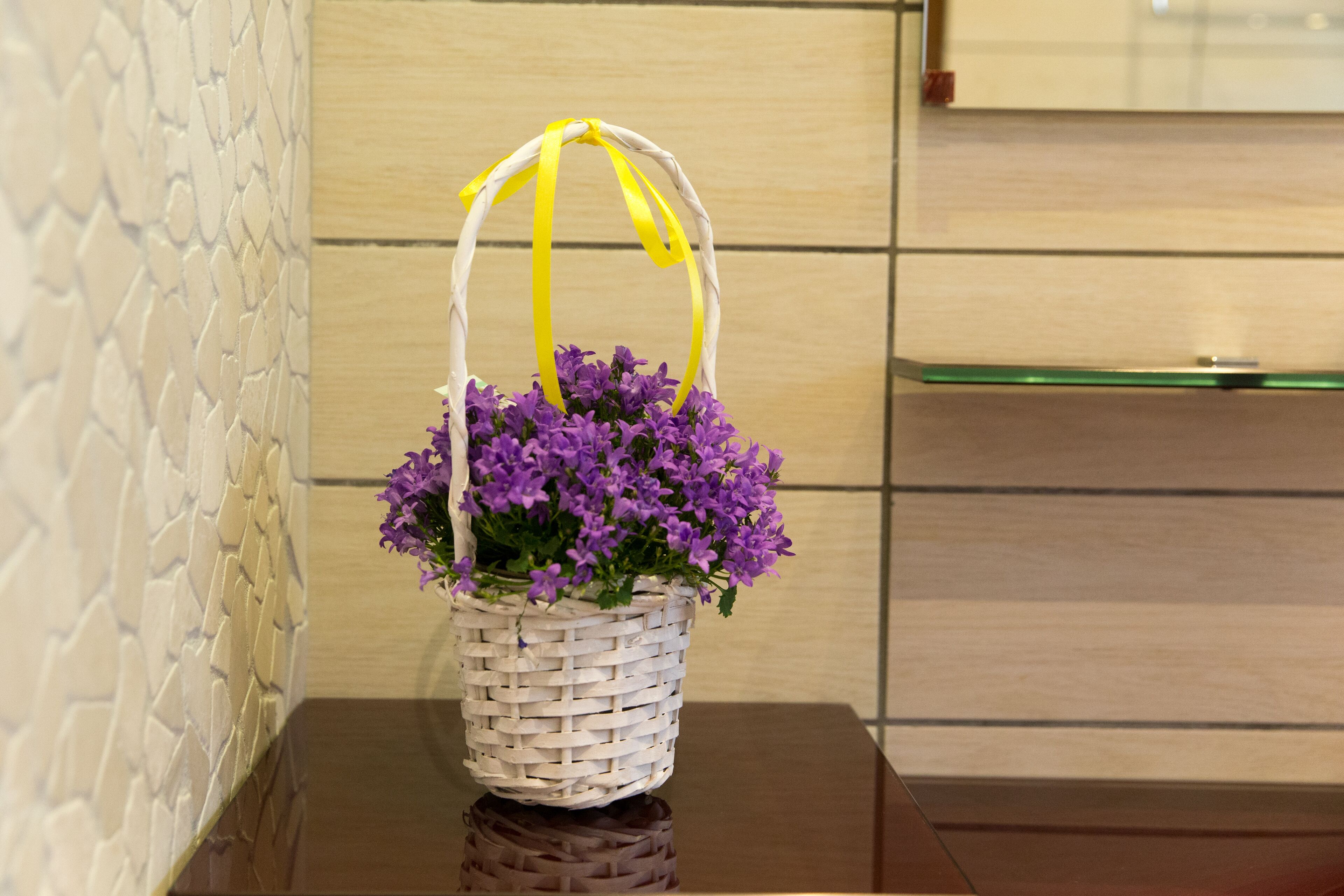 A white woven basket with purple flowers and a yellow handle on a reflective surface.