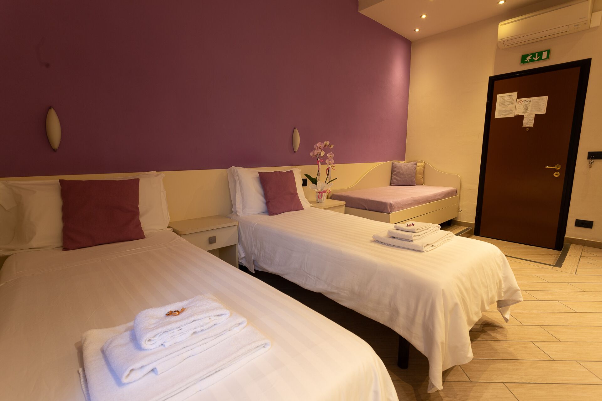 A hotel room with two beds, a small bench, and a door with an emergency exit sign.