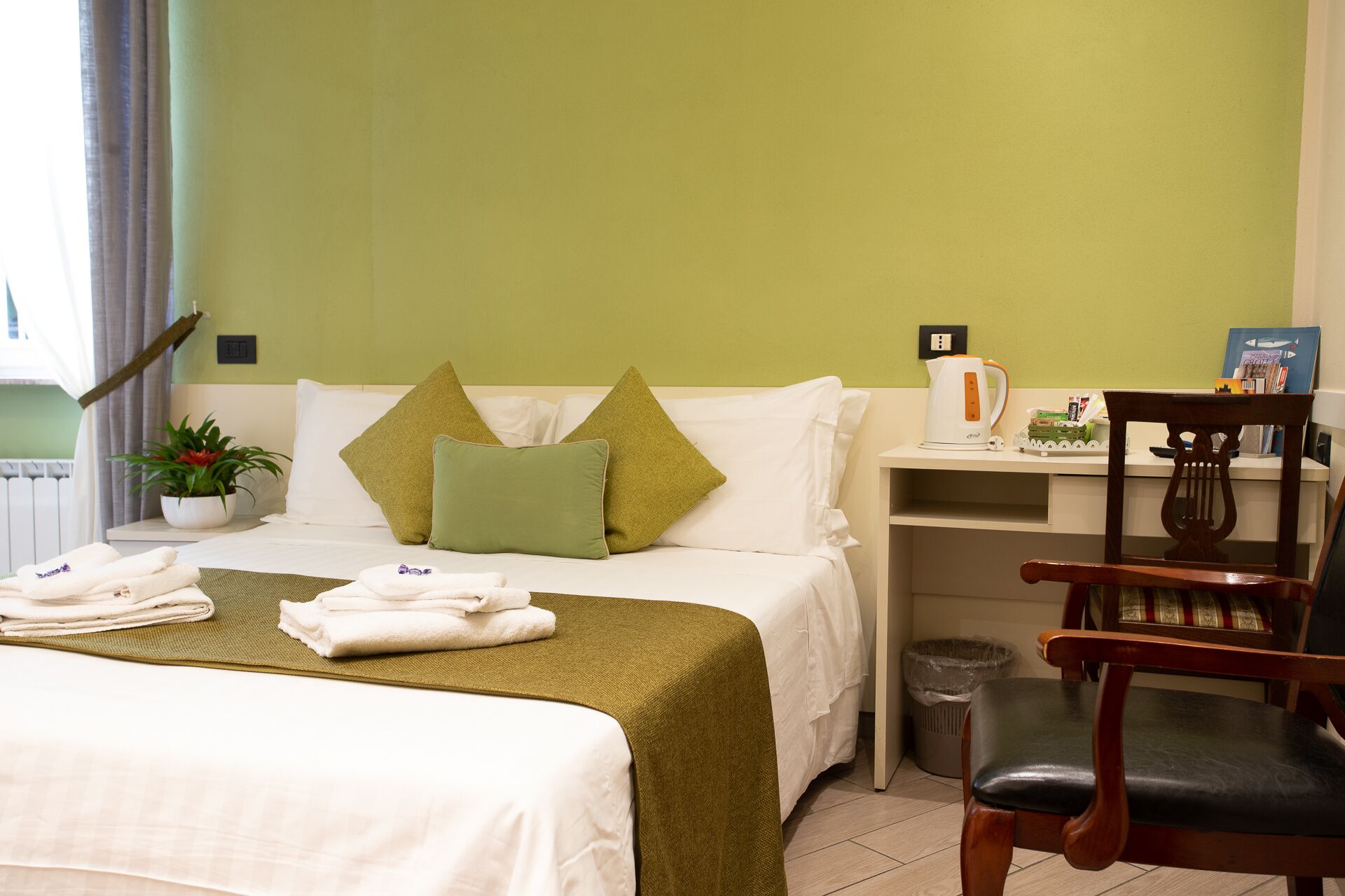 Ein Hotelzimmer mit Bett, Kissen, einem kleinen Tisch mit Wasserkocher, einem Sessel, einem Mülleimer und einer Zimmerpflanze.