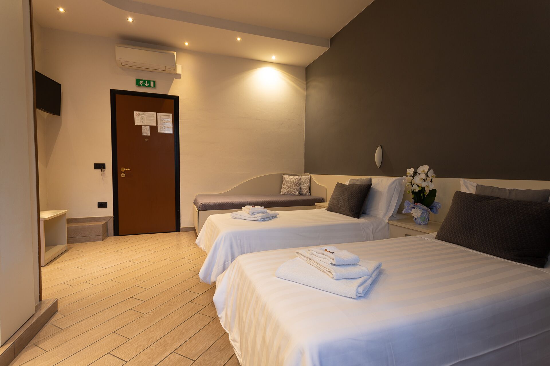 A hotel room with two beds, a small bedside table with a lamp, a wall-mounted TV, and a door with an emergency exit sign.