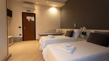 A hotel room with two beds, a small bedside table with a lamp, a wall-mounted TV, and a door with an emergency exit sign.