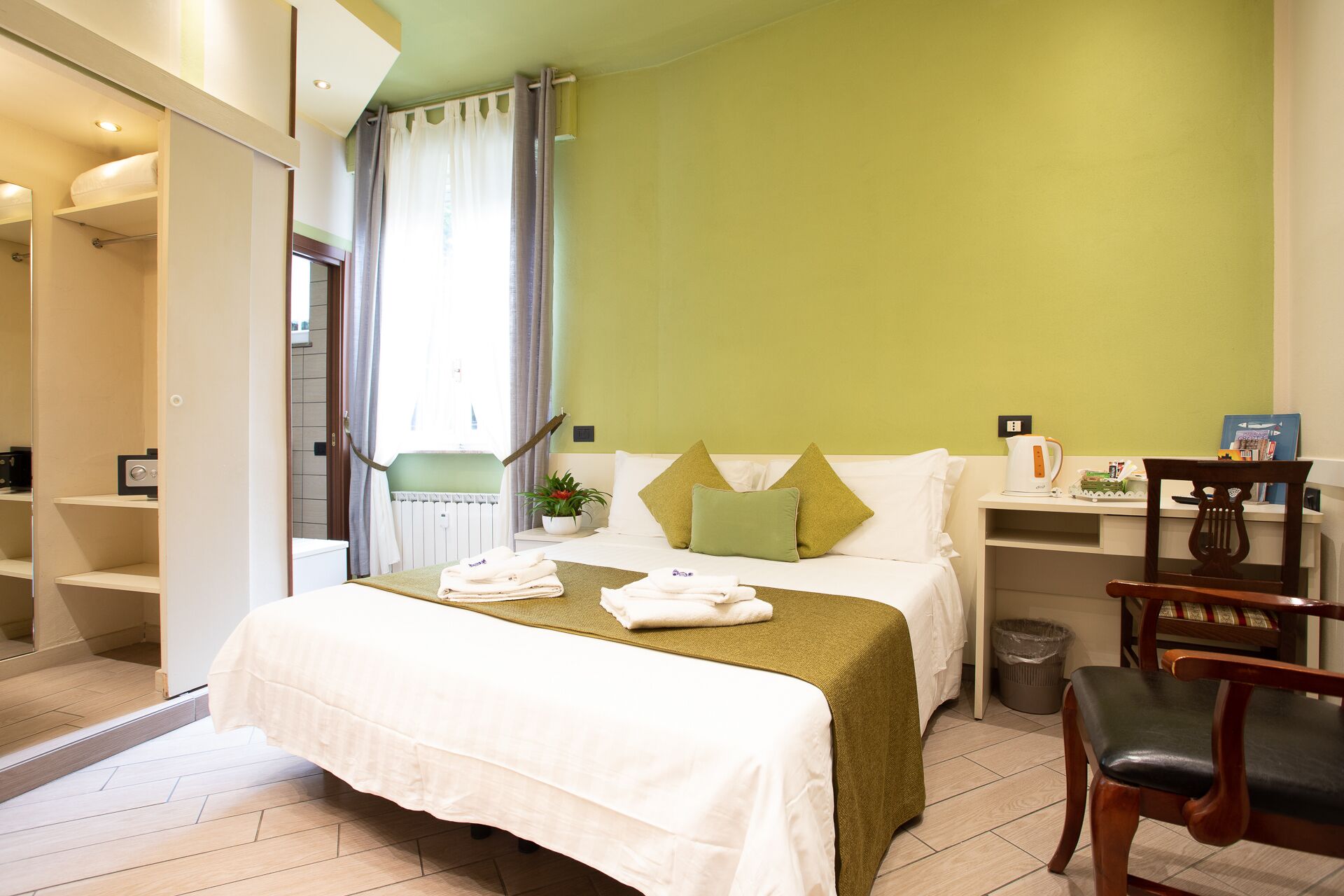 Una habitación de hotel con cama, escritorio, silla, radiador y ventana con cortinas.