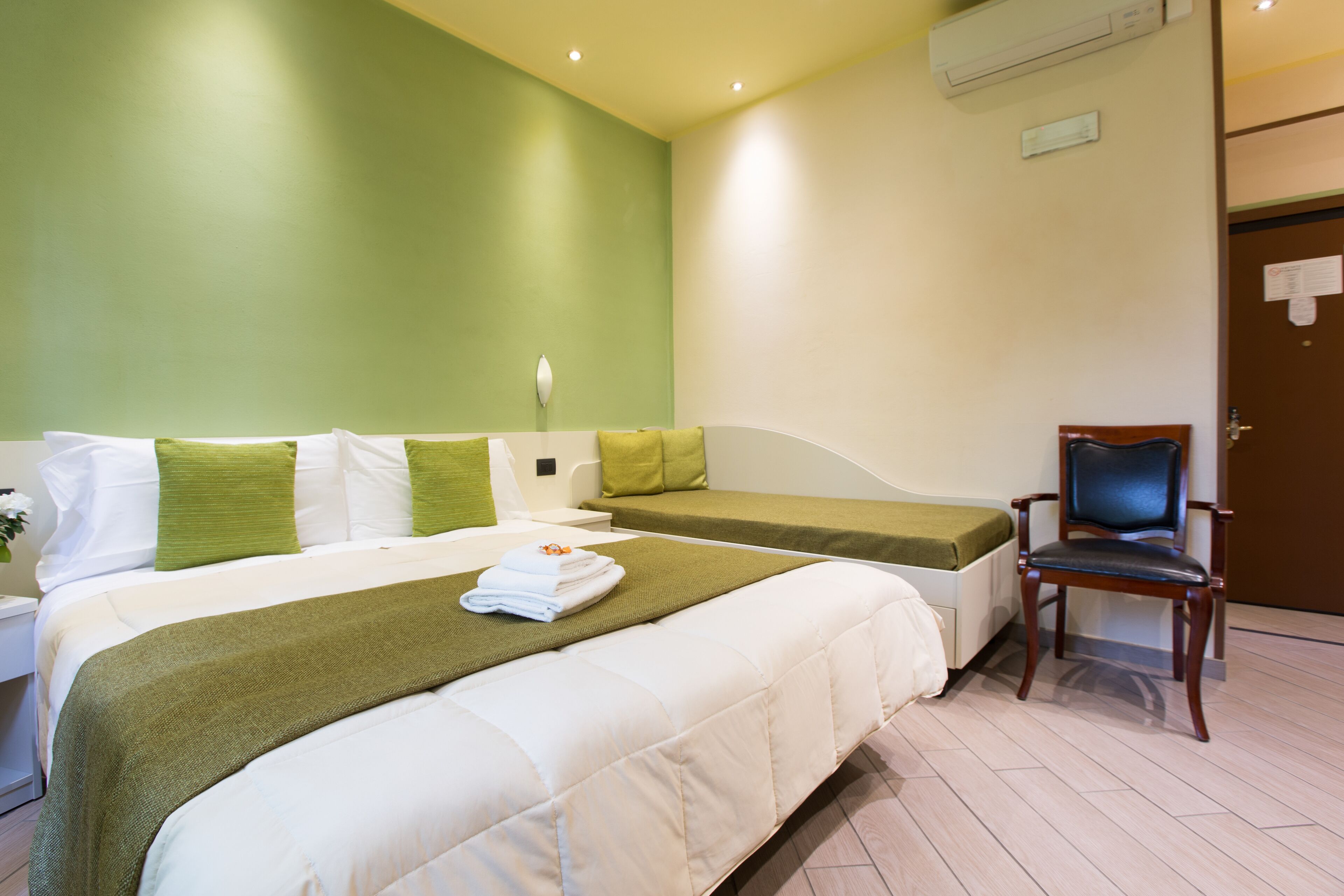 A hotel room with two beds, a chair, a nightstand, and a wall-mounted air conditioner.