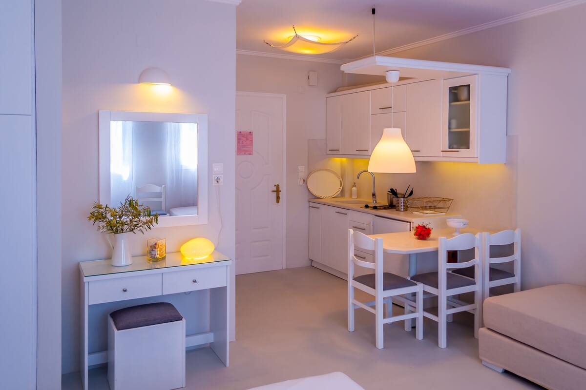 A compact kitchenette with white cabinets, a small dining table with chairs, a white sofa, and a mirror above the vanity.