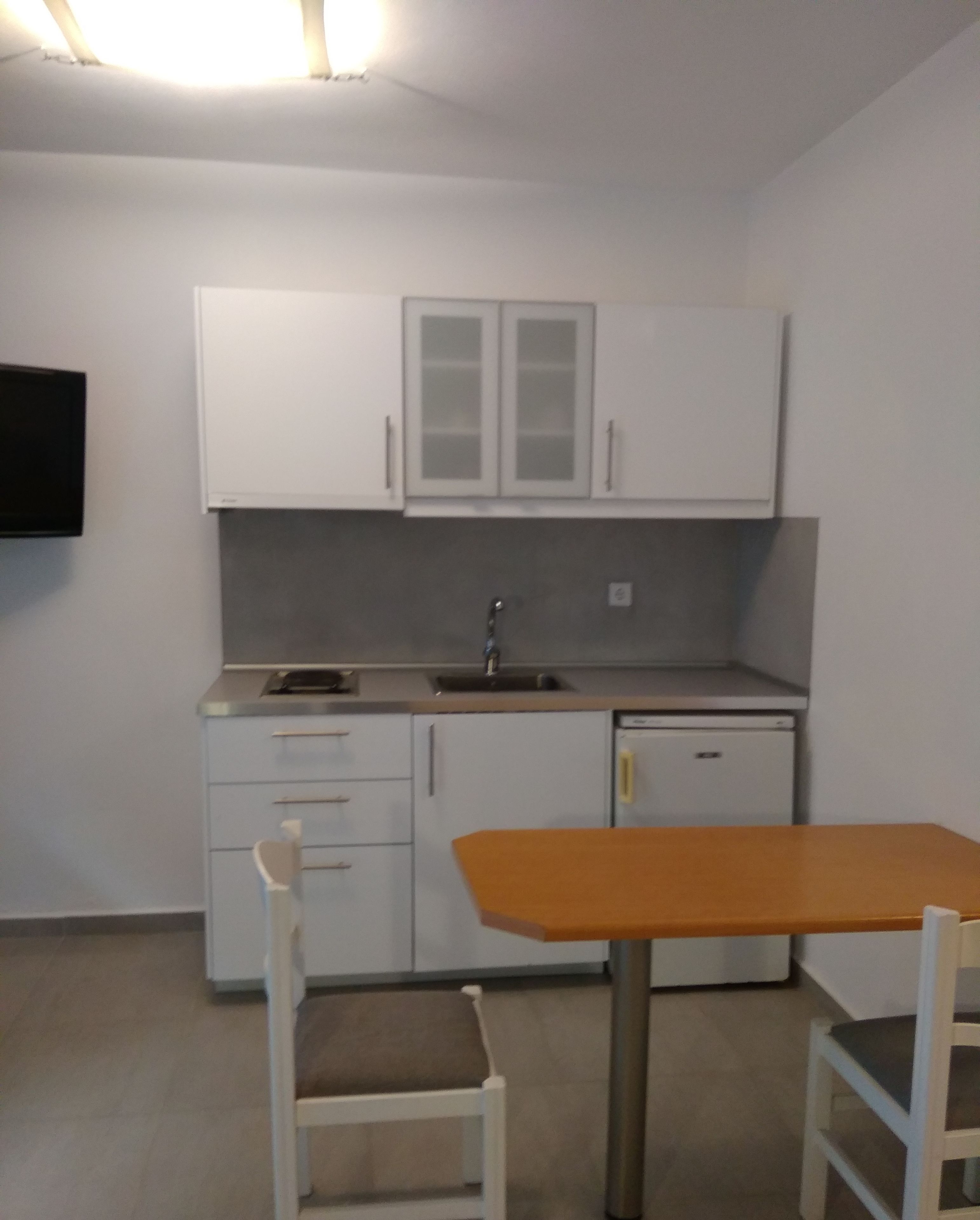 Een compacte keuken met witte kasten, een spoelbak en een kleine koelkast. Er staat een houten tafel met twee stoelen.