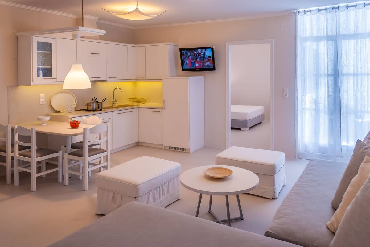 A modern, well-lit living room with a sofa, ottoman, and a small table. A television is mounted on the wall, and there is a kitchen area with white cabinets and a dining table.