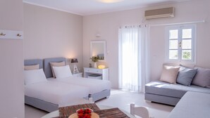 A modern bedroom with a bed, a sofa, a table with a fruit bowl, and a window with curtains. - Milos Bay Suites (Milos)