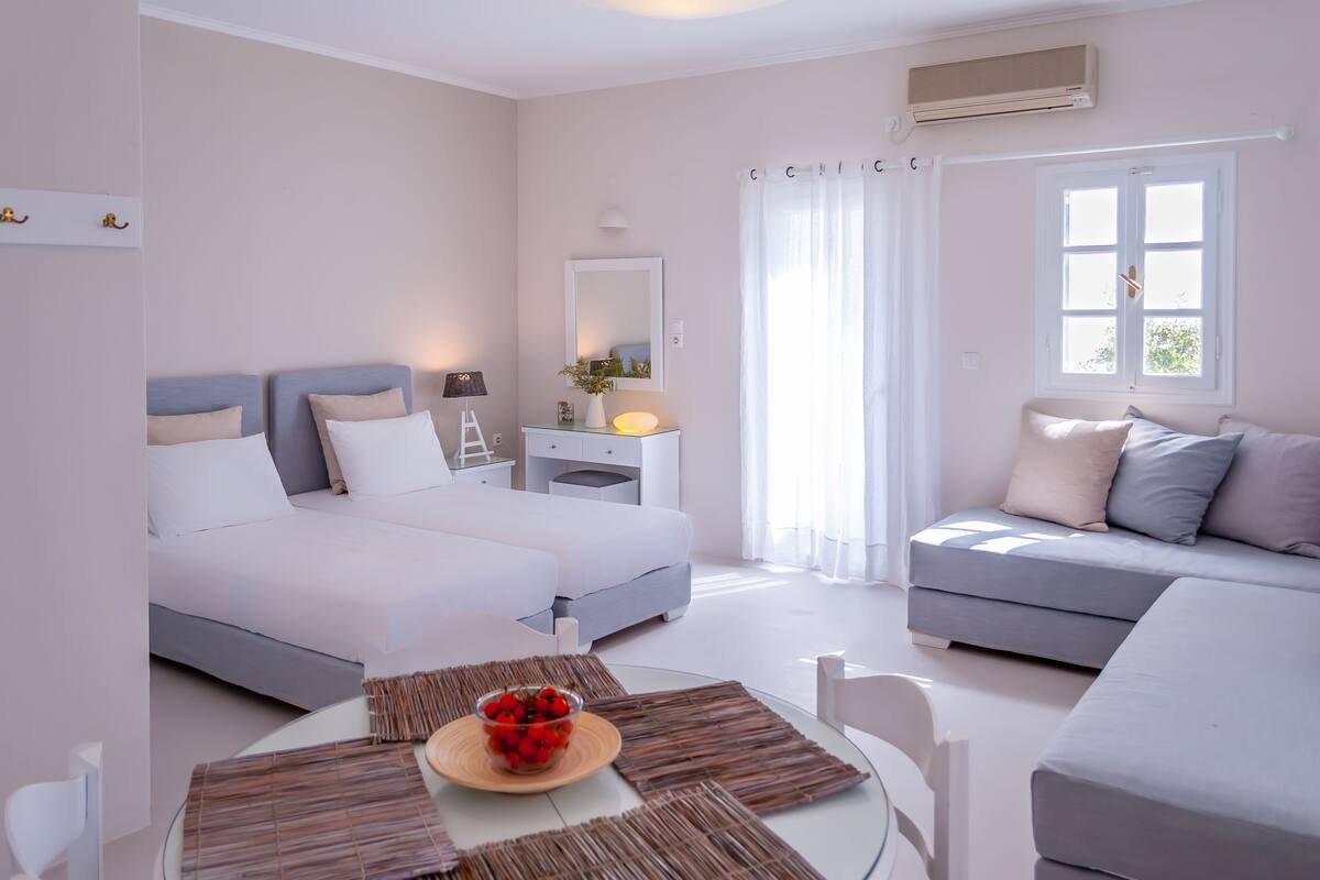 A modern bedroom with a bed, a sofa, a table with a fruit bowl, and a window with curtains.