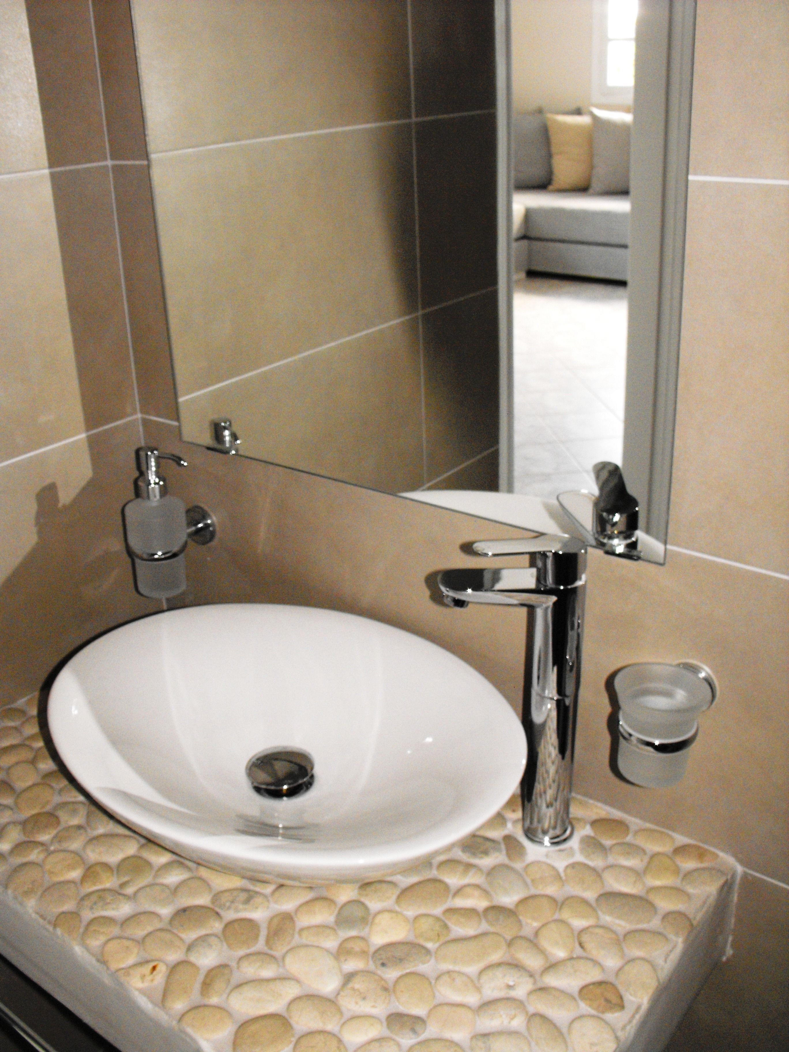 Une salle de bain avec un lavabo blanc, un robinet chromé et un miroir qui reflète un canapé et une fenêtre.