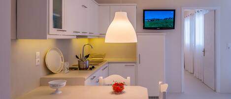 Una cocina moderna con armarios blancos, una mesa de comedor con sillas, un bol de fruta y un televisor en la pared.