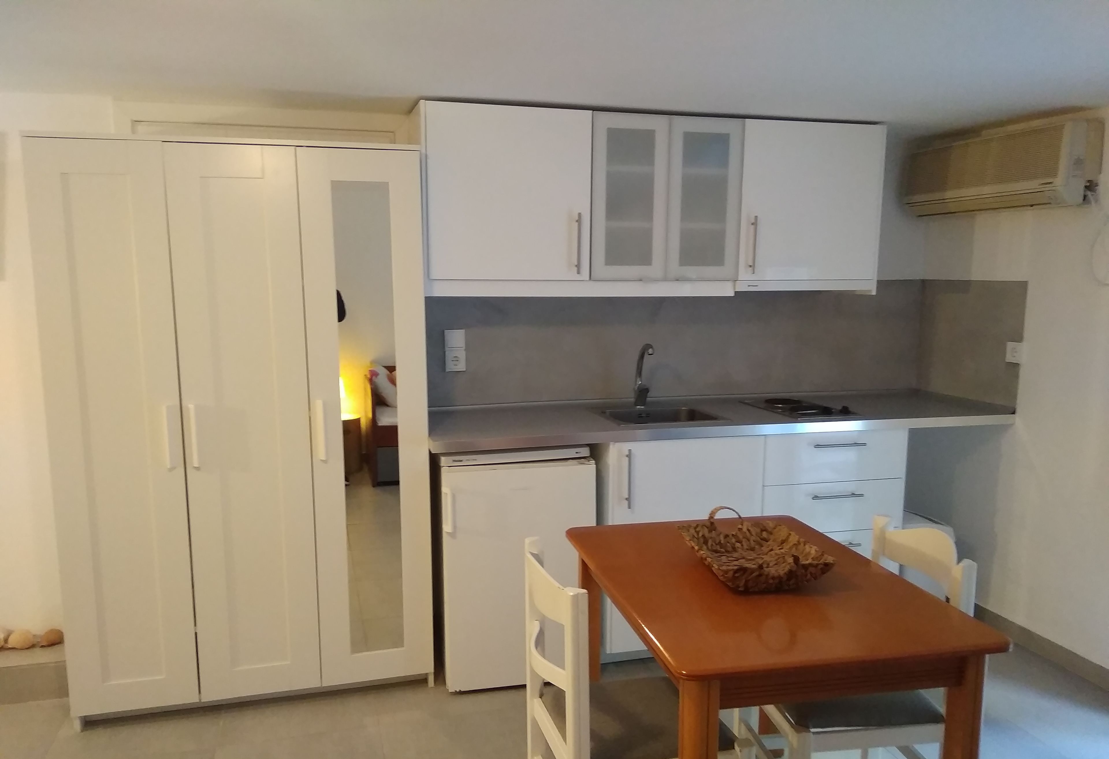 Una cocina compacta con armarios blancos, un refrigerador pequeño y una mesa de comedor con una cesta.