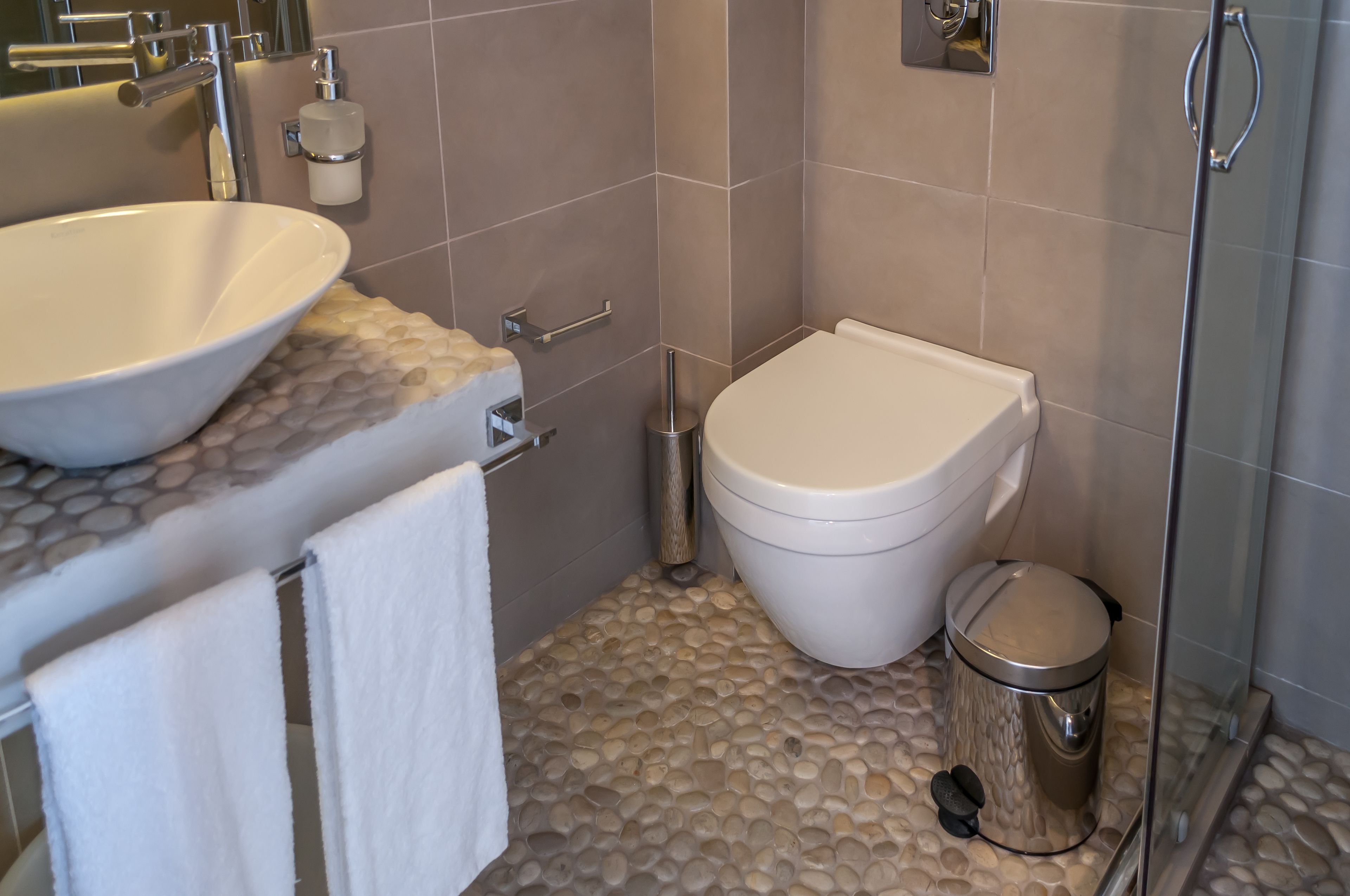 Un baño con lavamanos blanco, inodoro, y cubo de basura, suelo de baldosas y zona de ducha.