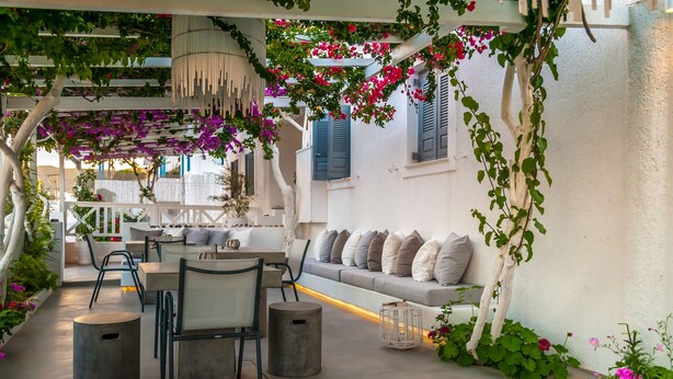A cozy outdoor seating area with a sofa and chairs, surrounded by lush greenery and flowering plants. - Milos Bay Suites (Milos)
