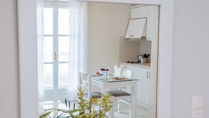 A modern kitchen with white cabinets, a dining table, and chairs. A large mirror reflects the room, and there is a vase with green plants on a white table. - Milos Bay Suites (Milos)