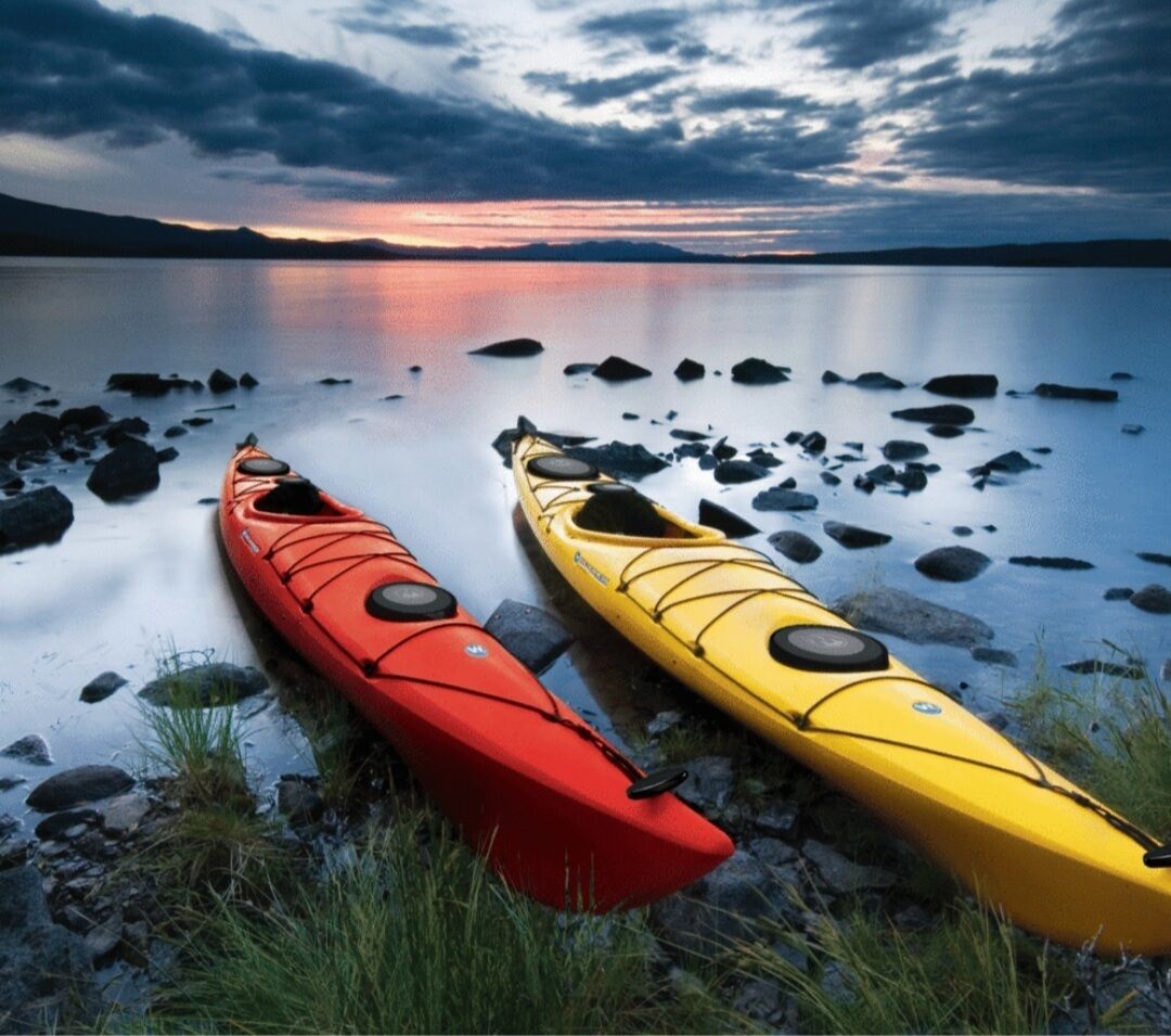 Two kayaks, one red and one yellow, on a rocky shore with calm water and a sunset in the background.
