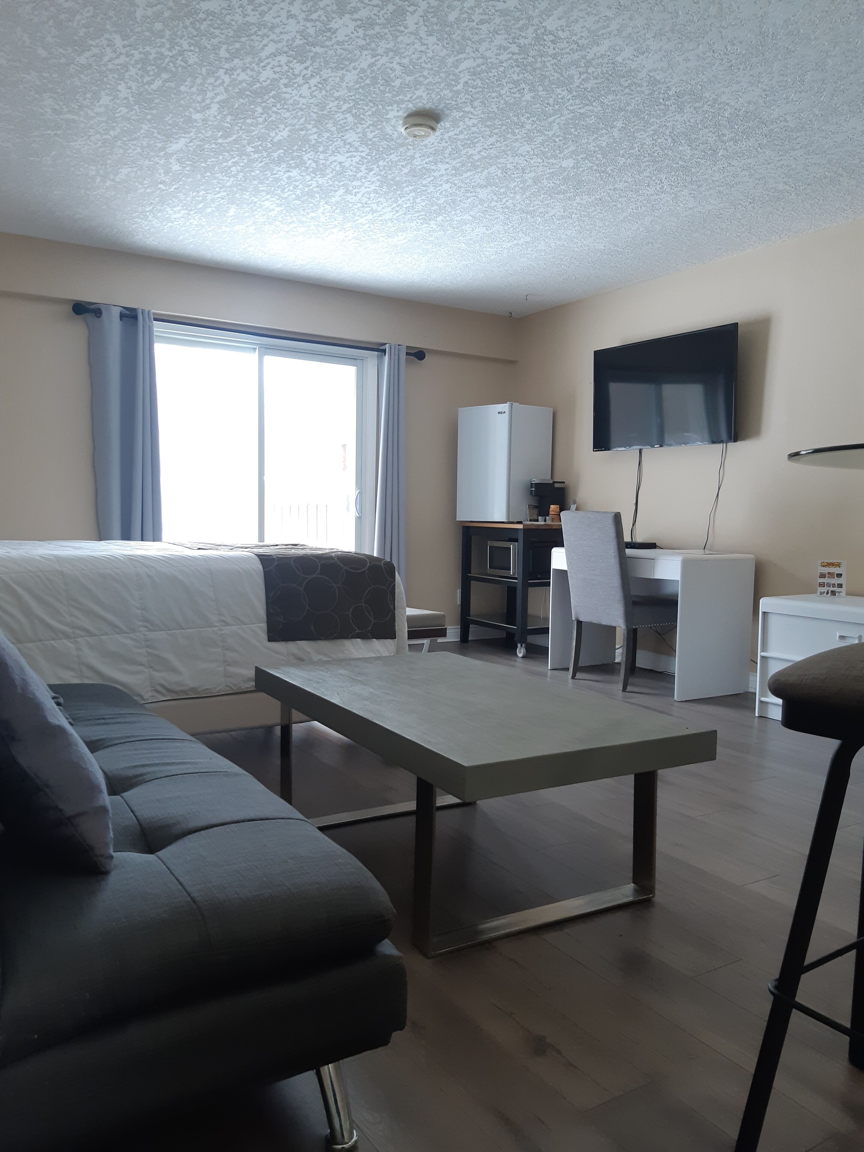 A modern hotel room with a bed, a sofa, a dining table, a TV, a refrigerator, and a desk.