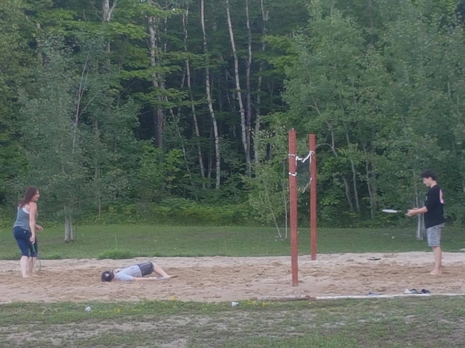 A sandy area with a person lying down, another person standing, and a third person holding a frisbee.