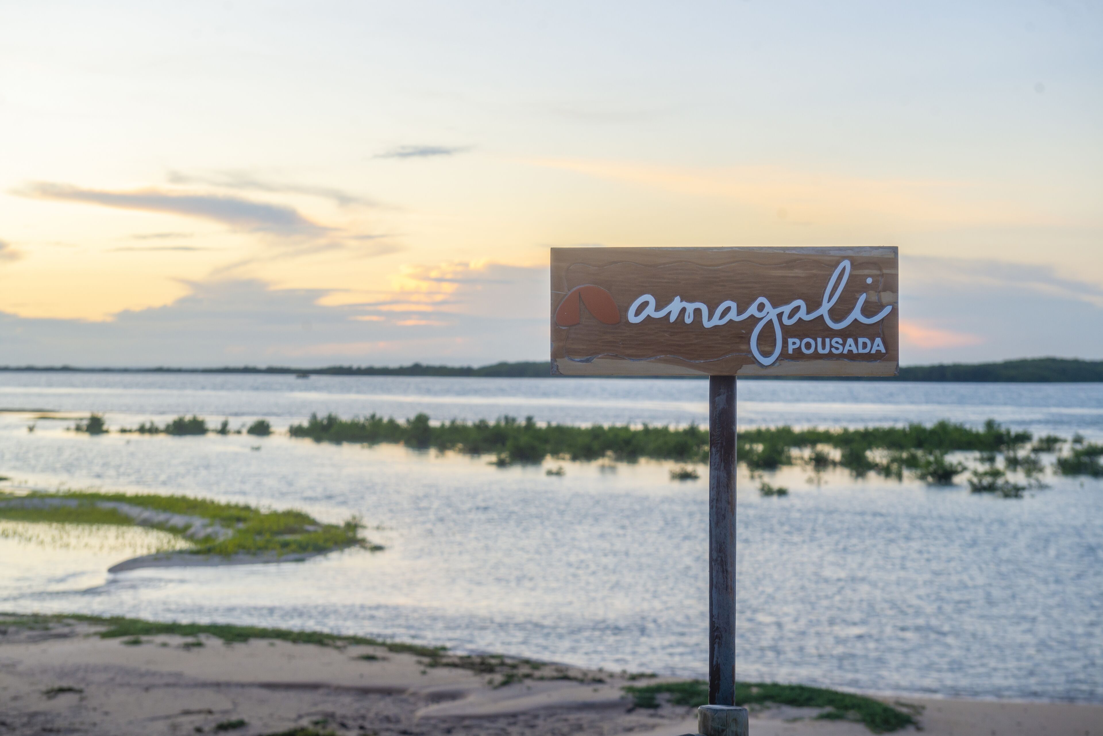 Un letrero de madera con el texto "amagali pousada" sobre un fondo de atardecer.