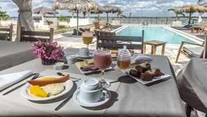 Taula preparada per esmorzar, amb fruita en un plat, un got de suc i una tassa de cafè, amb vista a una piscina i una platja.