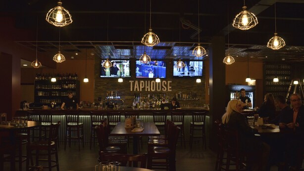 A bar area with multiple hanging light fixtures, a bar counter with a bartender, and several patrons seated at tables. - Rodeway Inn (Sudbury)