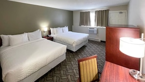 A hotel room with two beds, a desk, a chair, a lamp, an air conditioning unit, and a window with curtains. - Rodeway Inn (Sudbury)