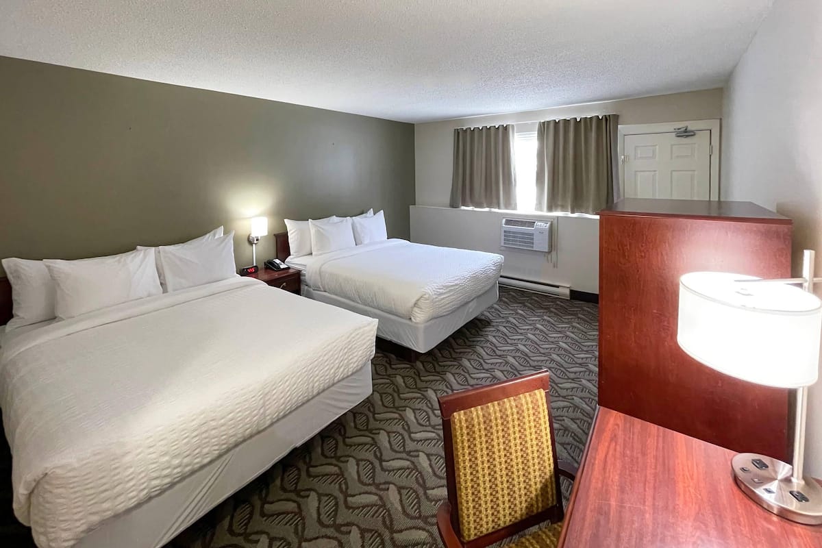 A hotel room with two beds, a desk, a chair, a lamp, an air conditioning unit, and a window with curtains.