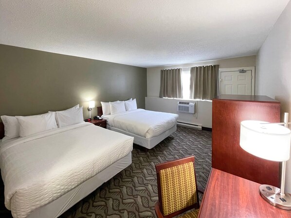 A hotel room with two beds, a desk, a chair, a lamp, an air conditioning unit, and a window with curtains.
