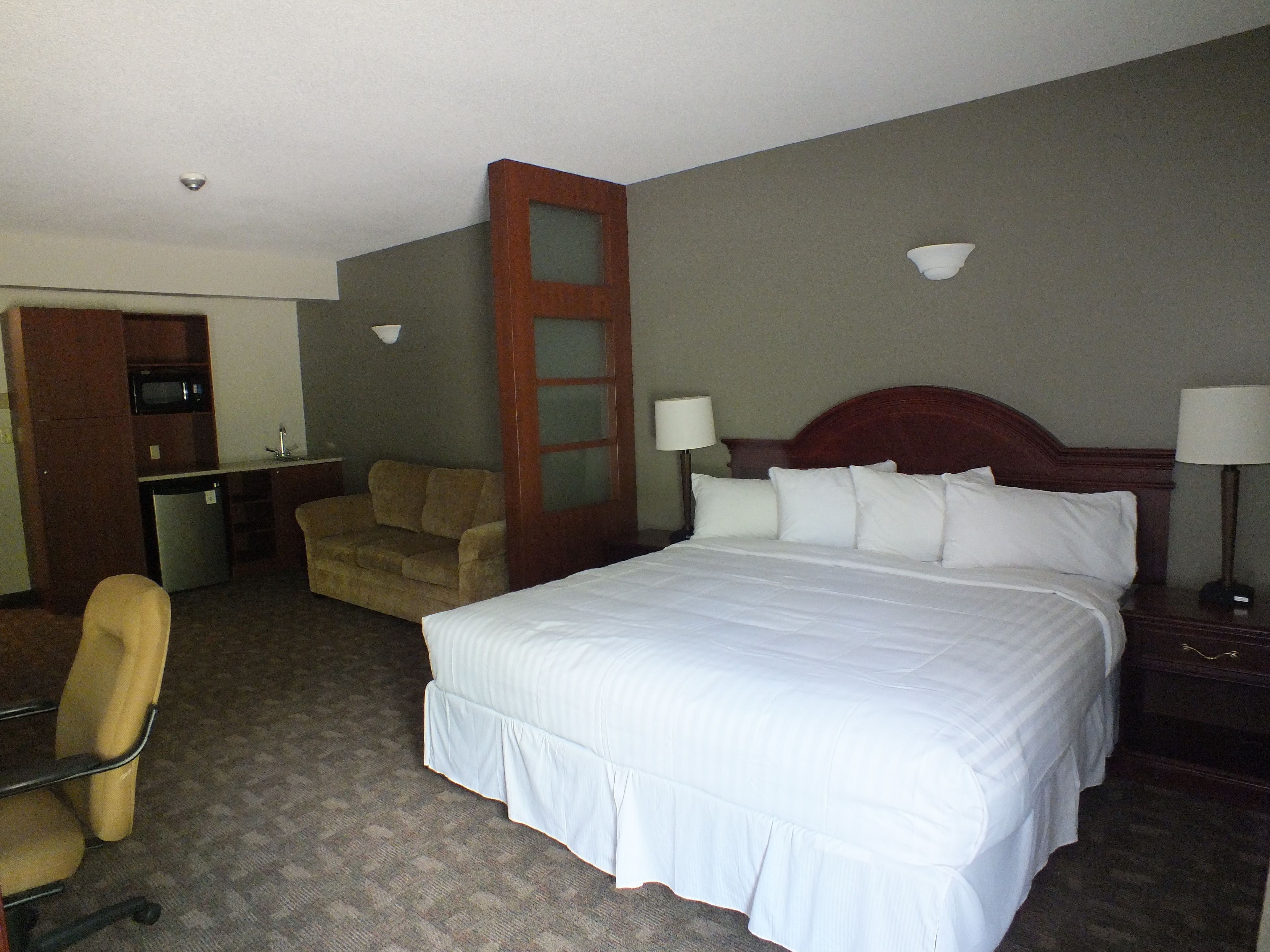 A hotel room with a large bed, a desk with a microwave, a chair, and a small kitchenette.