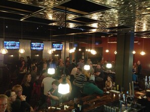 A crowded bar with hanging lights and multiple screens displaying content.