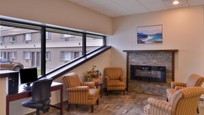 A modern office space with a desk, computer, and office chair, a fireplace with a stone surround, and a view of a building through a large window. - Rodeway Inn (Sudbury)