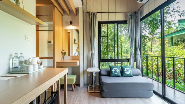 Une chambre moderne avec un bureau en bois, un canapé gris agrémenté de coussins, une petite table et une grande fenêtre donnant sur un paysage verdoyant.