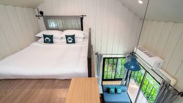 Een slaapkamer met een bed, een houten tafel, een bankje en een raam met uitzicht op groen.