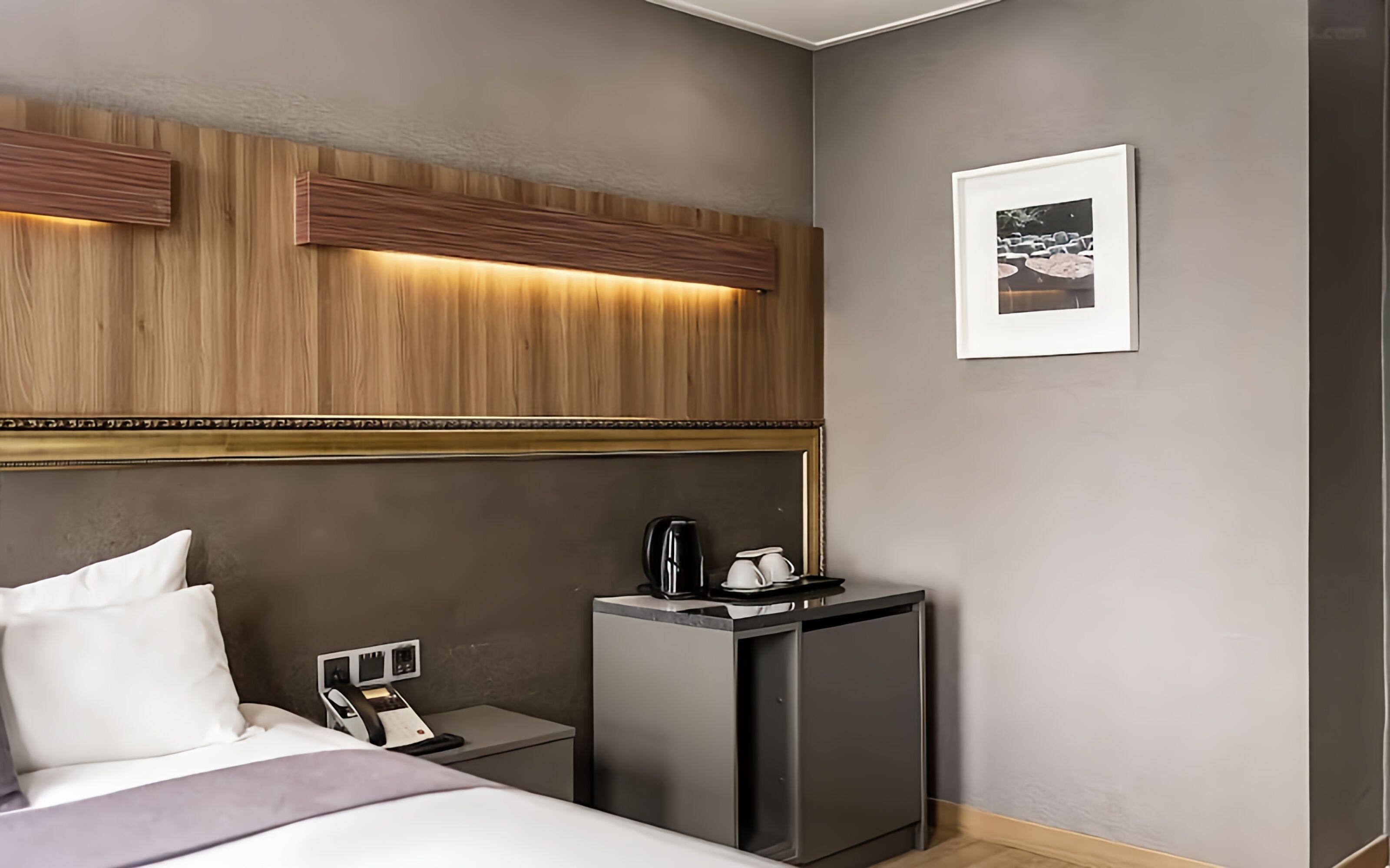 A hotel room with a bed, bedside table, a small desk with a coffee maker, a framed picture on the wall, and a wooden headboard with built-in lighting.