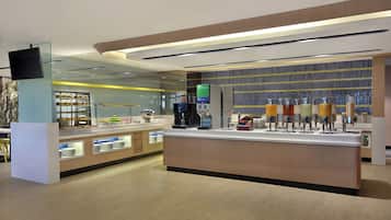 A modern cafe with a central counter, a coffee machine, and a variety of food items displayed.