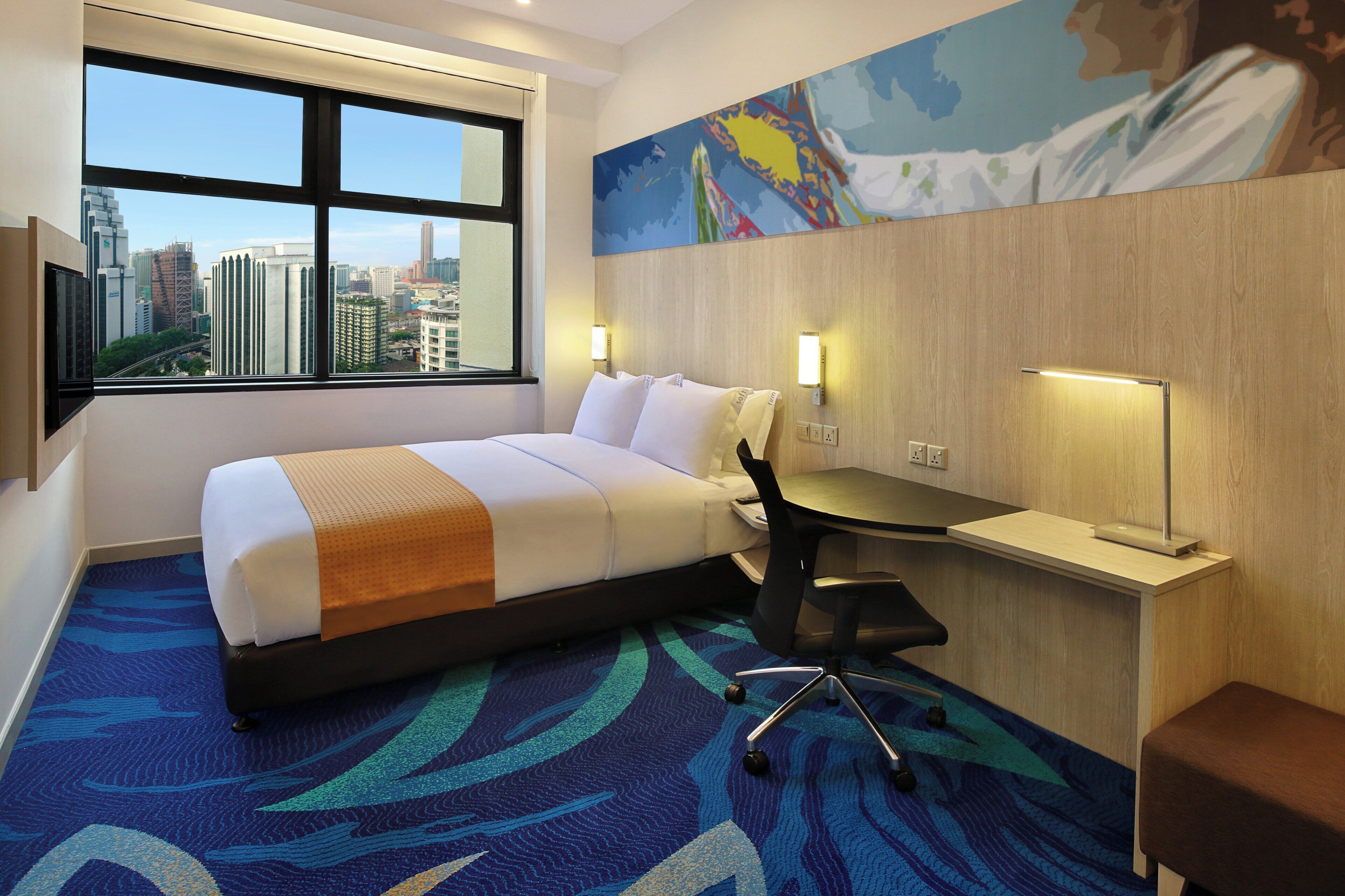 A hotel room with a bed, desk, chair, and a city view through large windows.