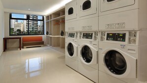 A laundry room with multiple washing machines and dryers, a bench, and a view of a cityscape through a window.