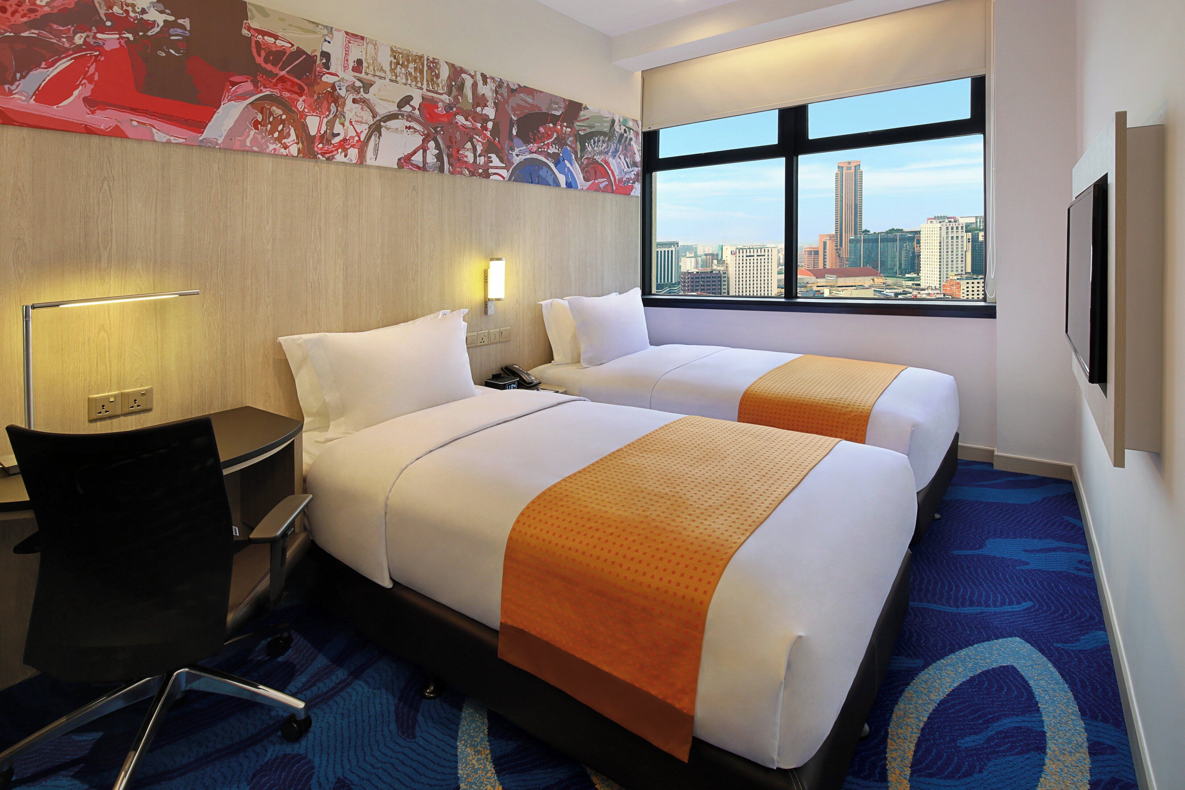 Habitación de hotel con dos camas, un escritorio, una silla, una lámpara, un ventanal con vista a la ciudad y un cuadro abstracto grande en la pared.