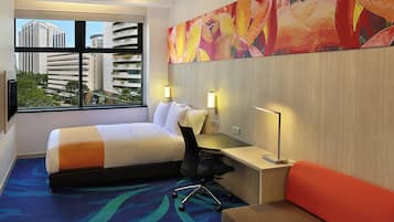 A hotel room with a bed, desk, chair, and a large window offering a city view.