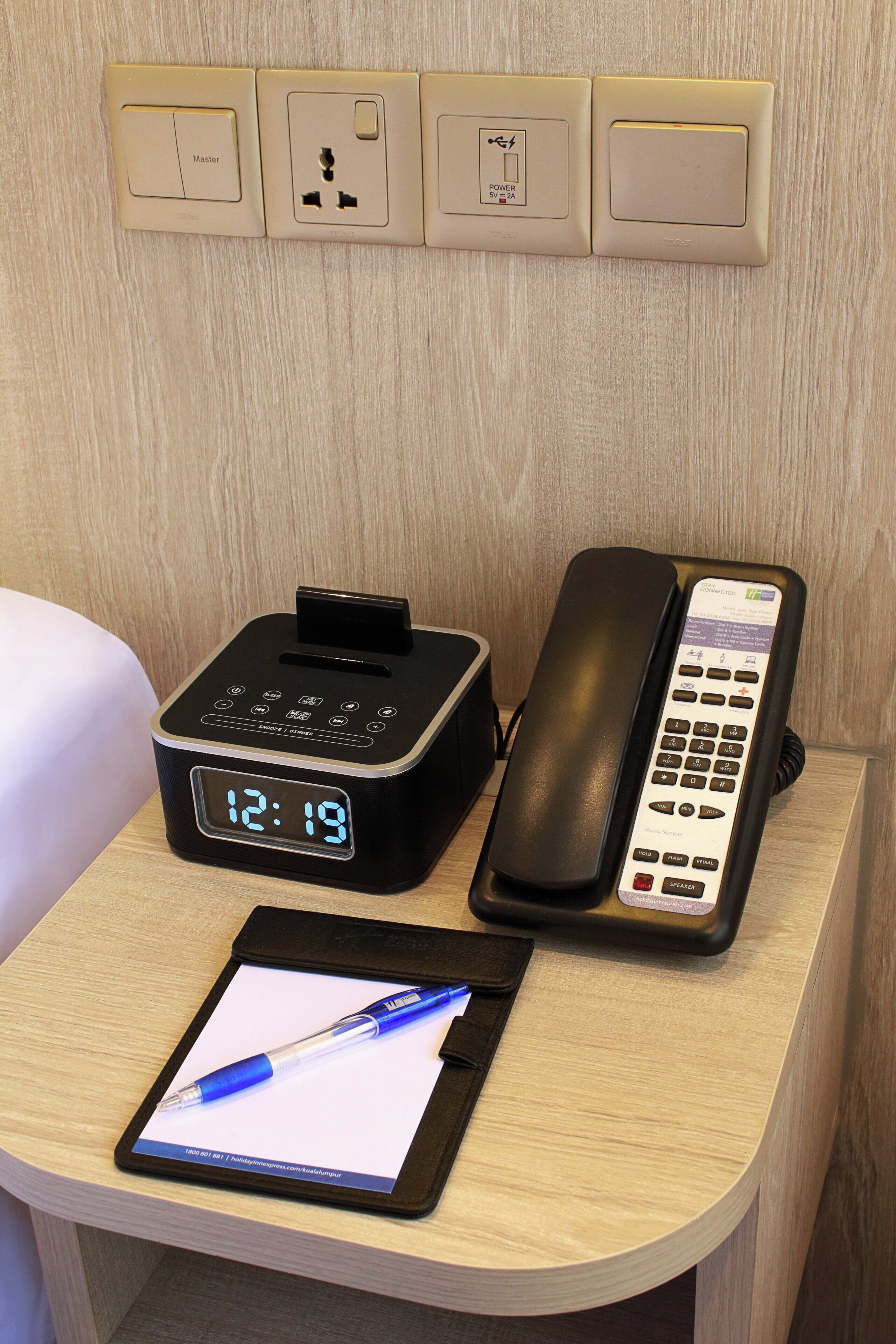 Een hotelkamer met een nachtkastje, voorzien van een digitaal klokje, een telefoon en een tablet.