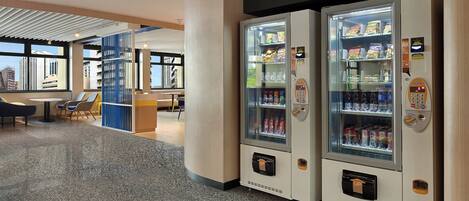 A modern hotel lobby with a seating area, a vending machine, and a view of the cityscape.