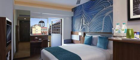 Habitación de hotel con cama, escritorio, silla, ventilador de techo y vista a edificios.