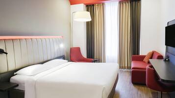 Habitación de hotel con una cama grande, un escritorio con silla, un sofá rojo, un televisor y una ventana con cortinas.