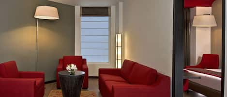 A modern hotel room with red sofas, a round table with a vase, a floor lamp, and a large window with blinds.