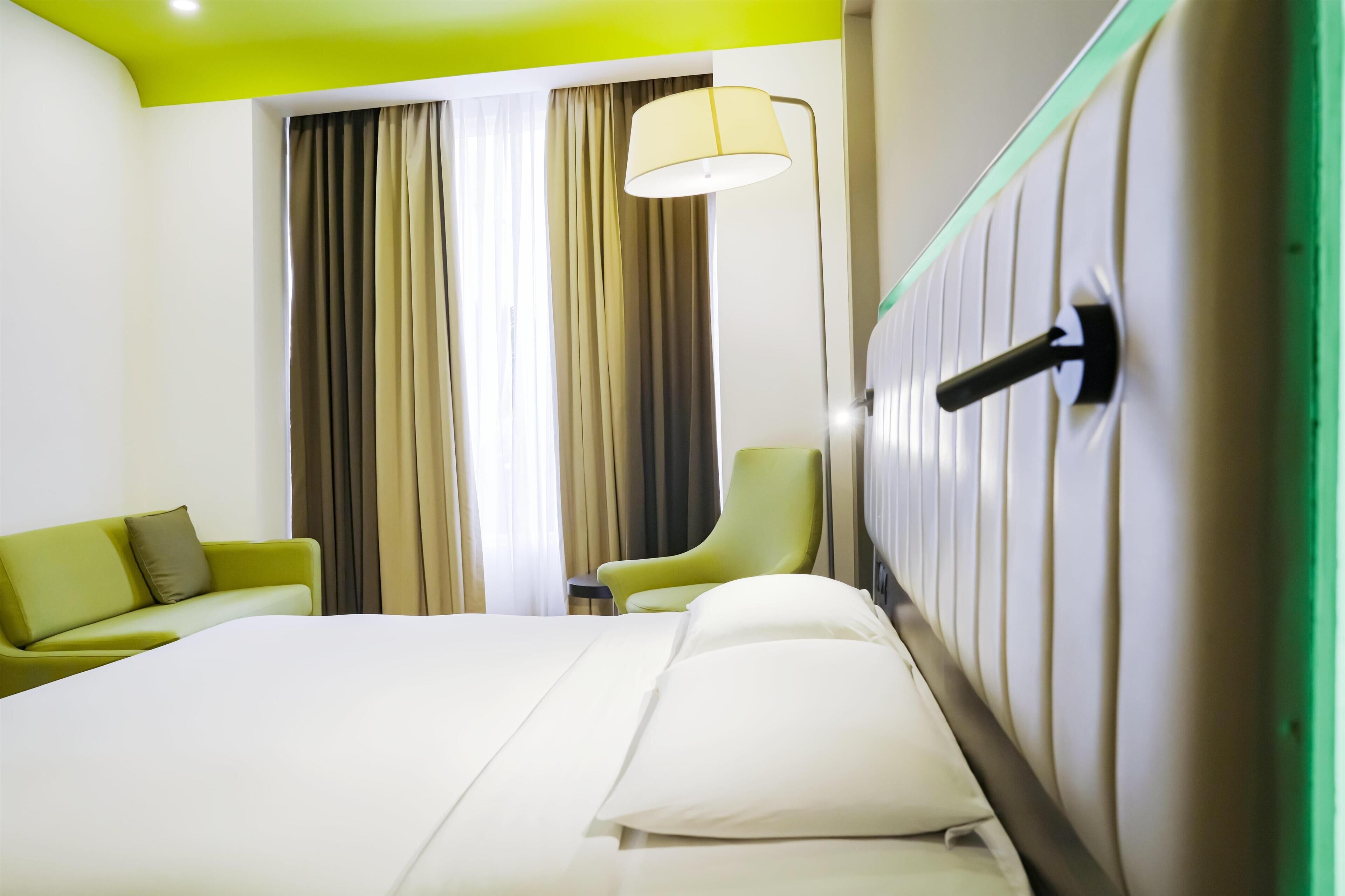 A hotel room with a bed, a green armchair, a sofa, a lamp, and a window with curtains.