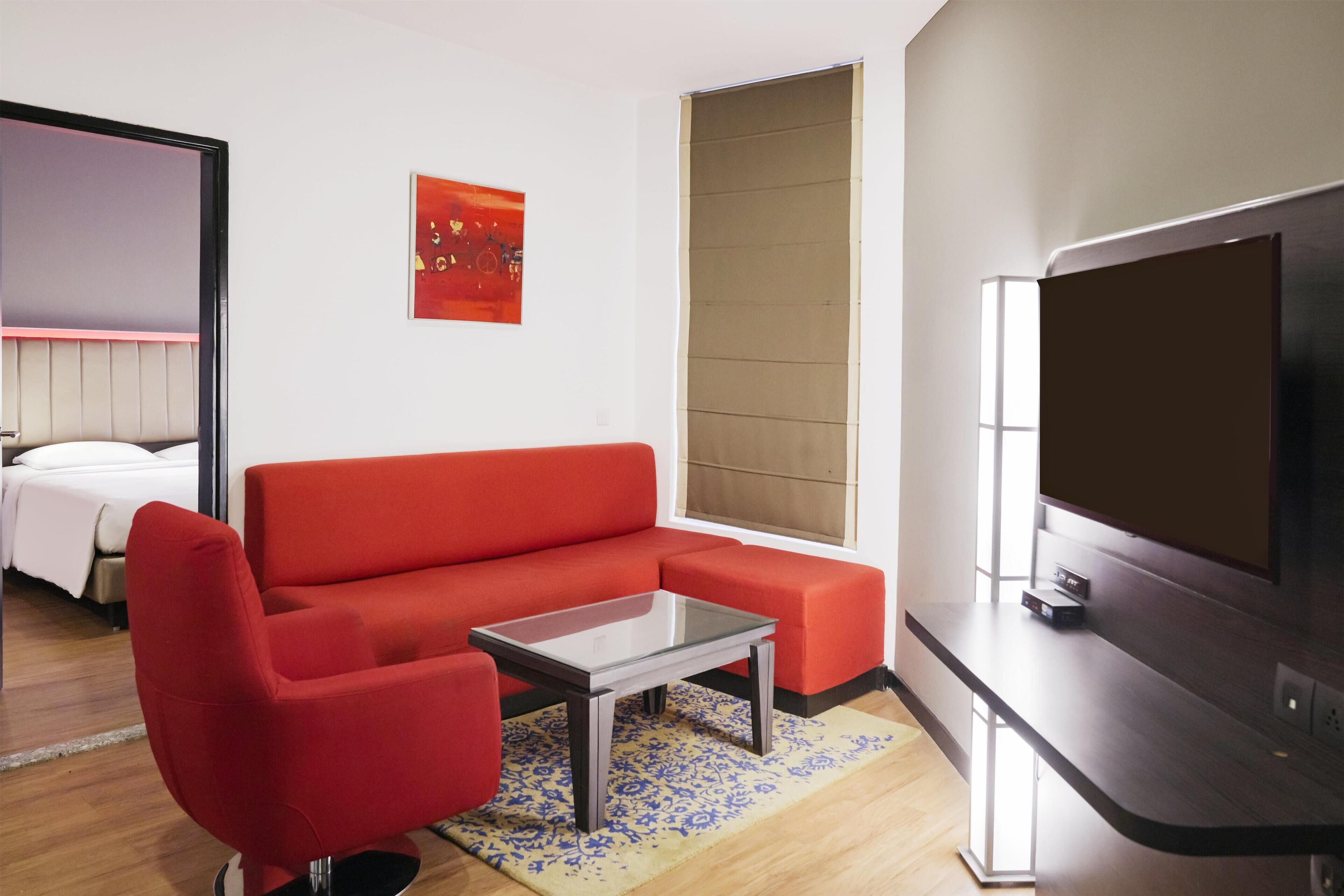Ein modernes Zimmer mit einem roten Sofa, einem Glastisch, einem Flachbildfernseher und einem Bett mit weißer Bettwäsche.