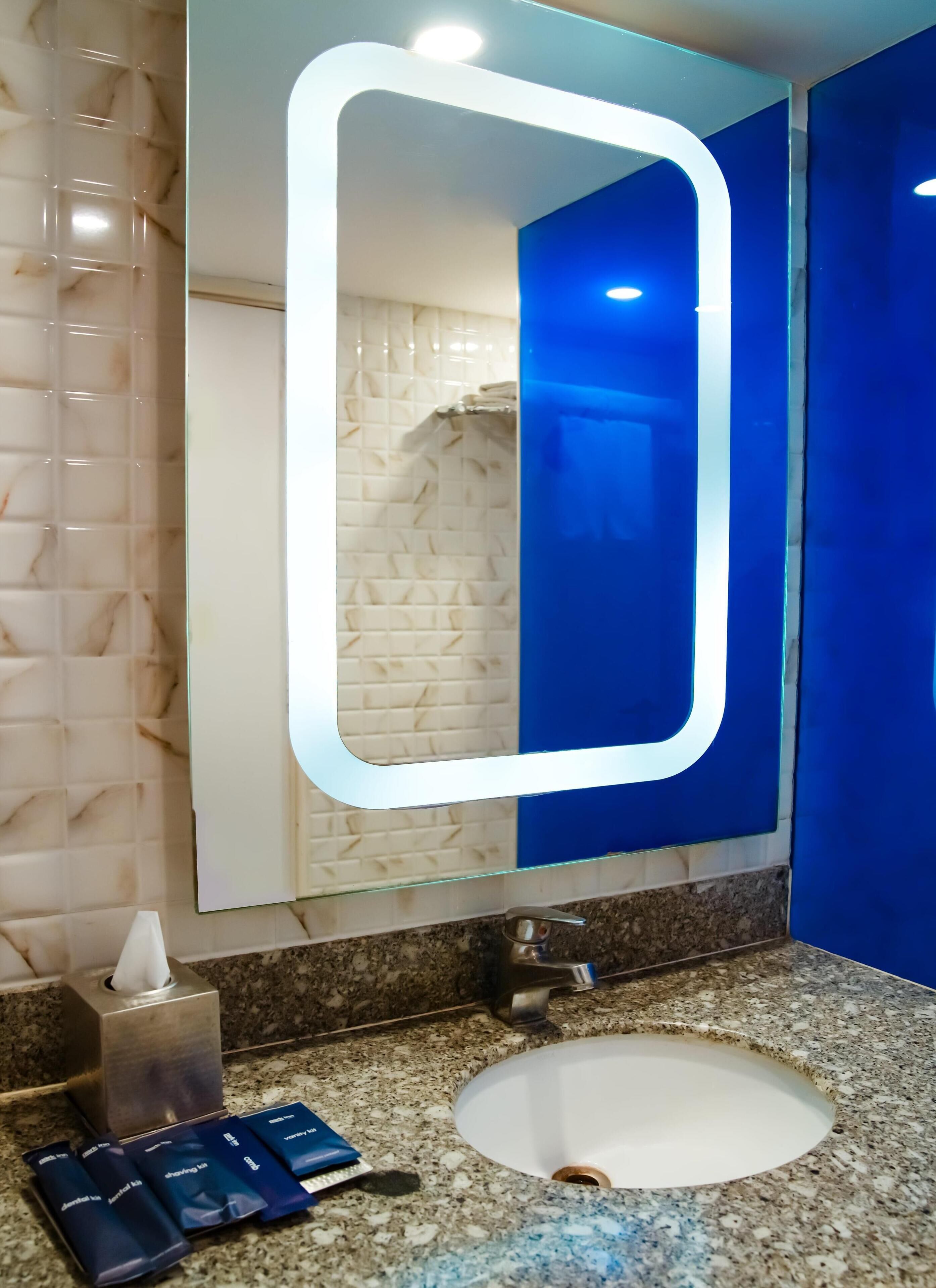 A bathroom with a marble wall, a sink with a granite countertop, a tissue dispenser, and a mirror with a light.