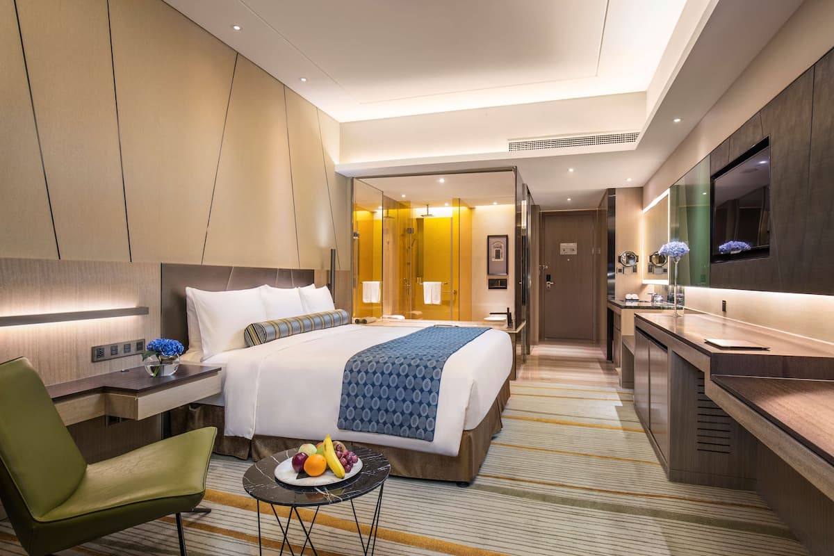 A hotel room with a bed, bedside table, chair, and a small table with fruit.