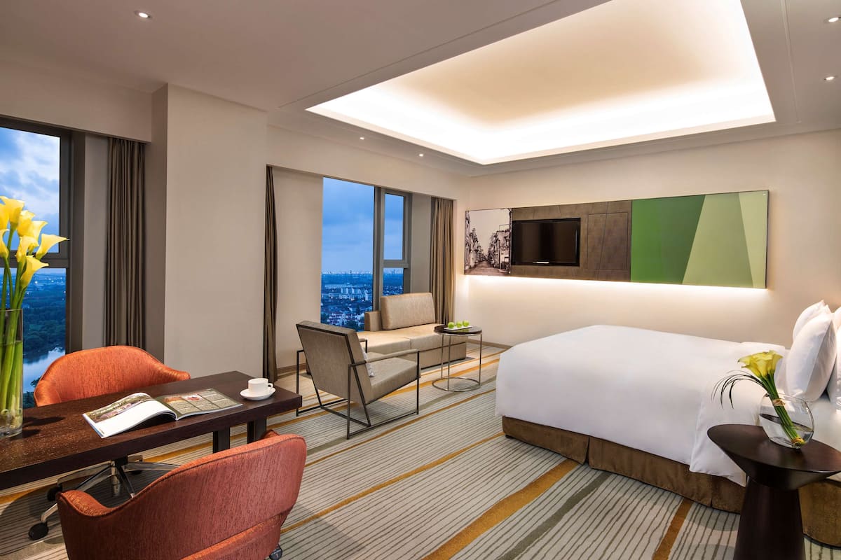 A modern hotel room with a large bed, a desk with a chair, a sofa, a TV mounted on the wall, and a view of the cityscape through large windows.